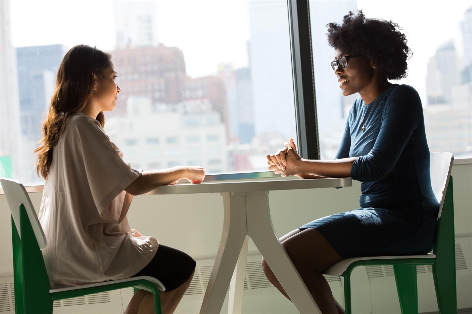 Two women of colour sitting at a table in front of a window, talking. 