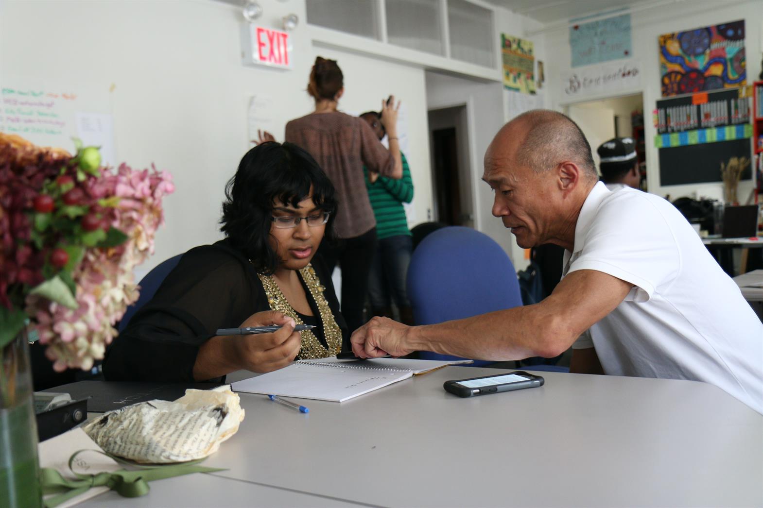 A teacher and student sitting at a table, the teacher is pointing to a workbook on the table and explaining something to the student