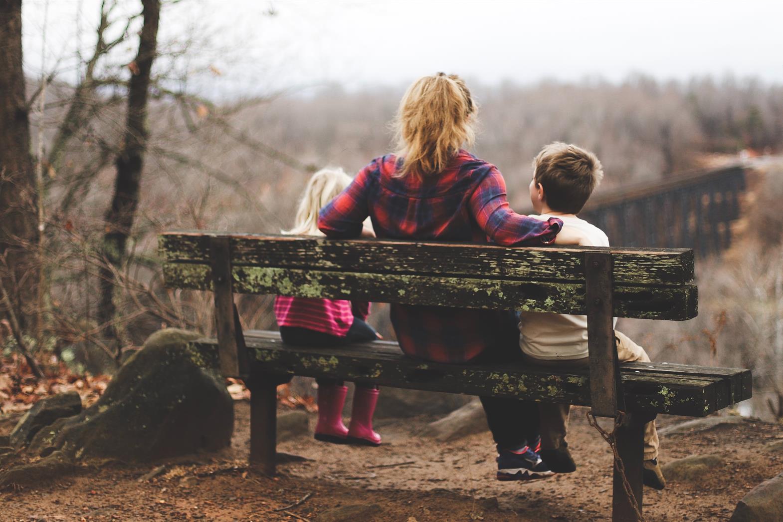 a mother and her two children sit on a bench on a cloudy day. They look out over a pretty valley in the autumn