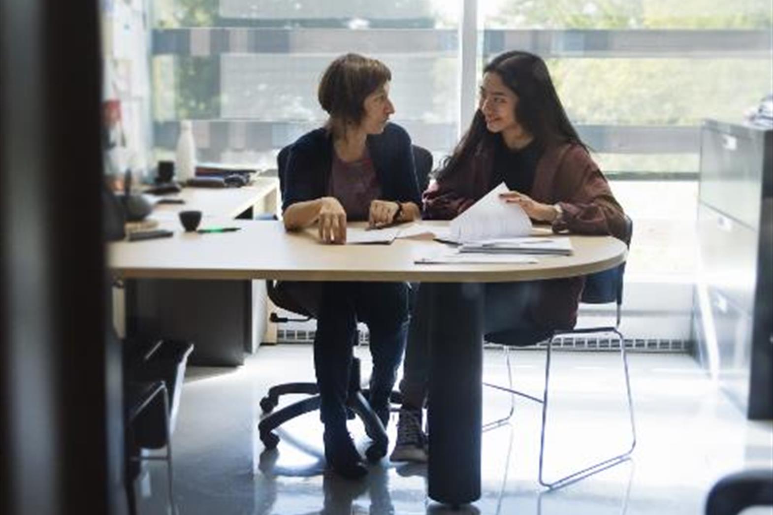 Two women sitting at a desk talking and looking at some papers. 