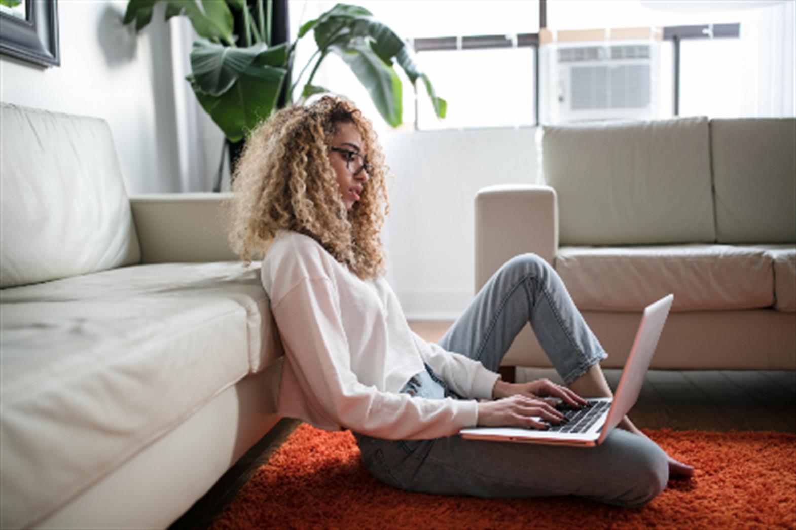 a teenager sitting on the floor, working on a laptop that is balanced on her leg. 