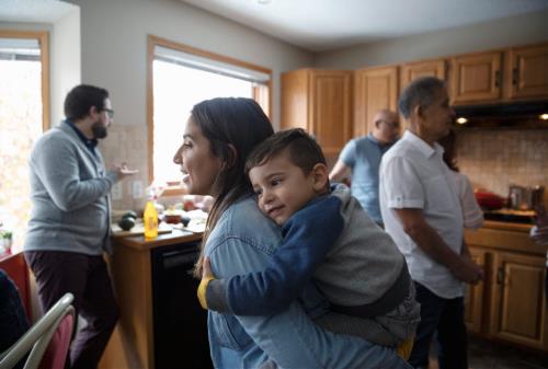 several people in a kitchen. In the foreground is a mother with a young child riding on her back piggy back style. They are both smiling