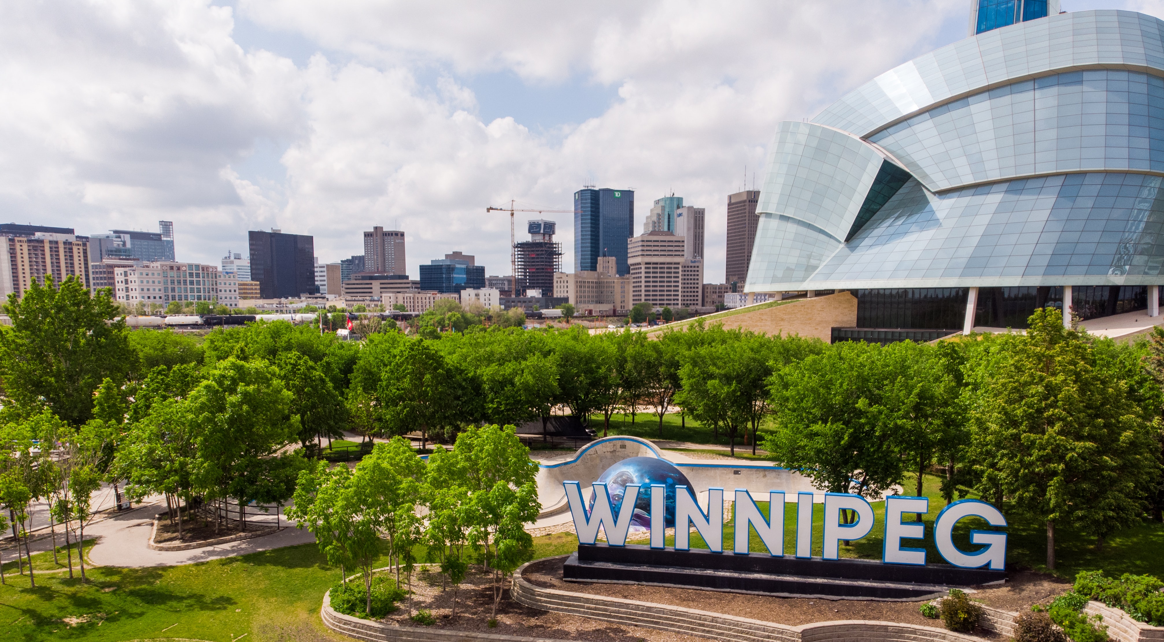 A sign in front of a blue and white building. The sign saws Winnipeg. There are green trees and bushes around the sign.