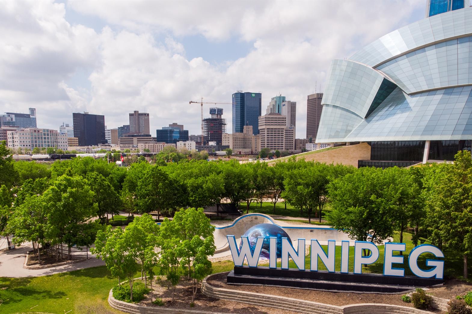 A sign in front of a blue and white building. The sign saws Winnipeg. There are green trees and bushes around the sign.